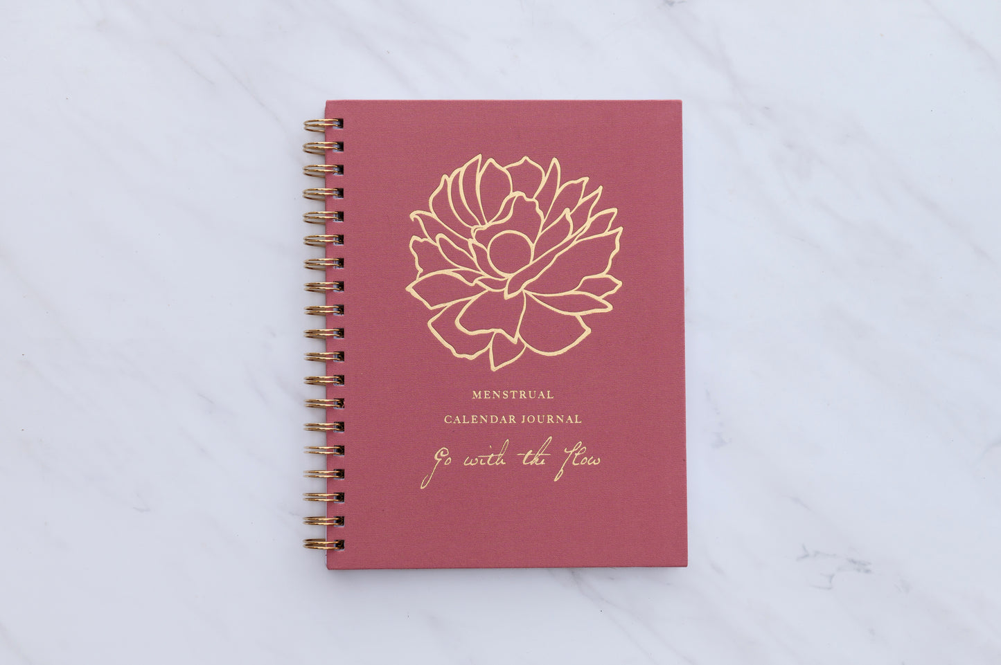 A red menstrual calendar journal with a floral design and the title 'Menstrual Calendar Journal: Go with the Flow' on the cover, which is placed on a white marble surface.